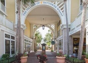 A covered walkway with a sign reading 'The Village of Baytowne Wharf' flanked by buildings with windows and palm trees.