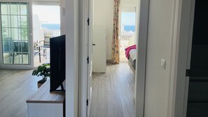 Une pièce avec un téléviseur posé sur un meuble, une plante en pot et une vue sur la mer, visible à travers une porte-fenêtre coulissante.