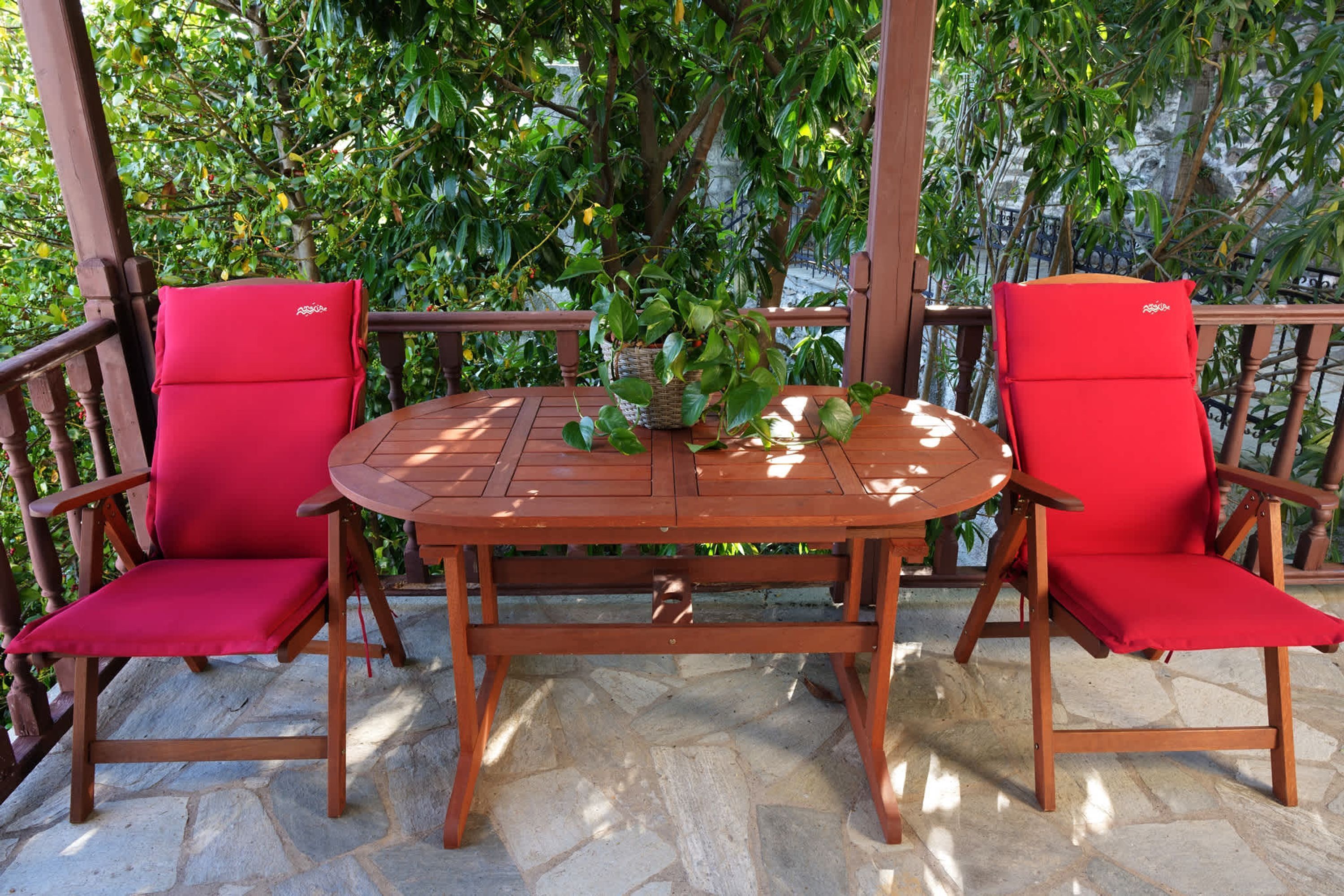 A wooden table and chairs with red cushions, set on a stone patio with a view of lush green trees.