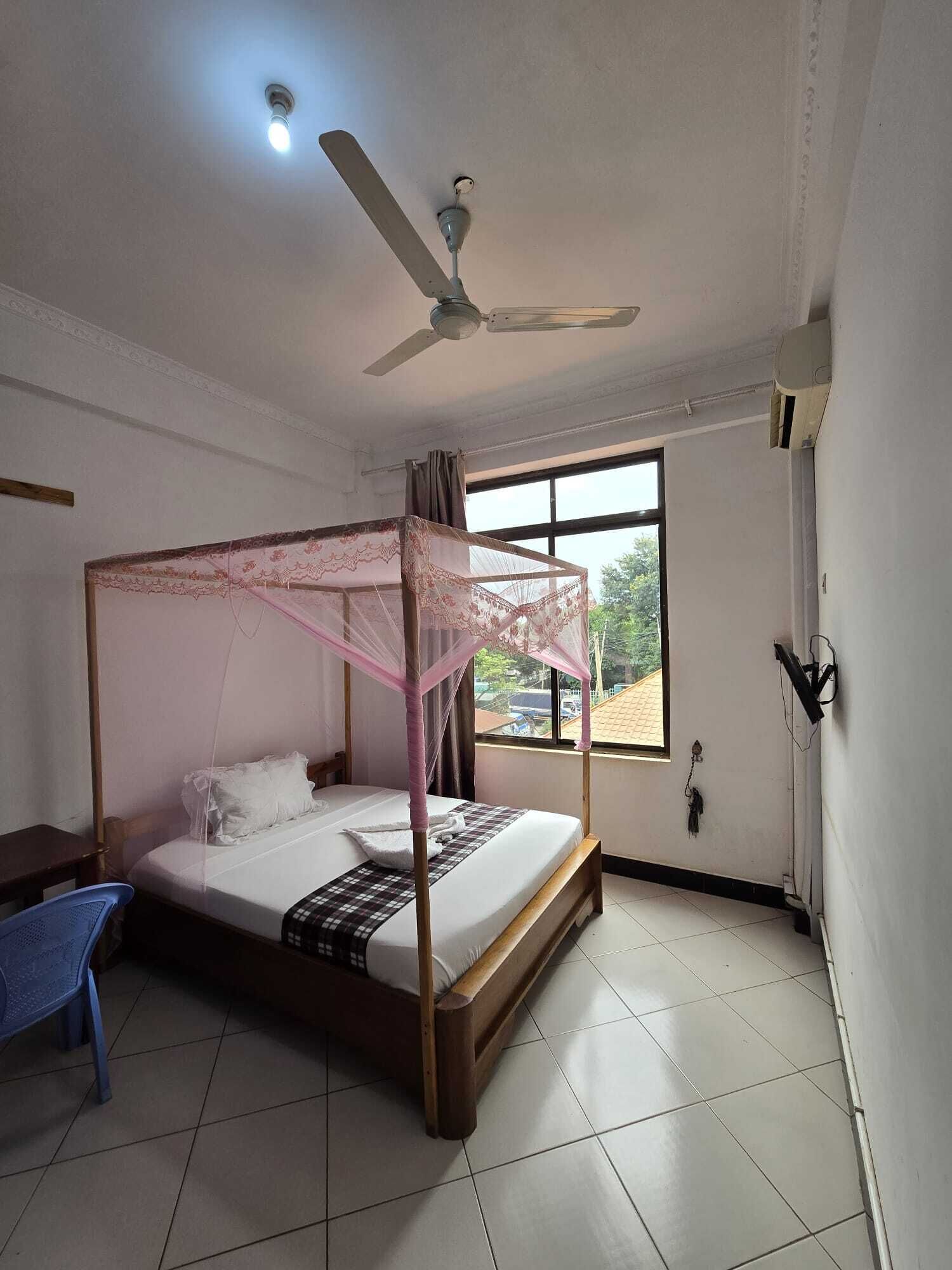 A single bed with a canopy, a ceiling fan, a window with a view of the outside, and a blue chair.