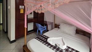 Un lit simple avec un baldaquin, un bureau, une chaise et un ventilateur de plafond dans une pièce aux murs blancs et au sol carrelé.