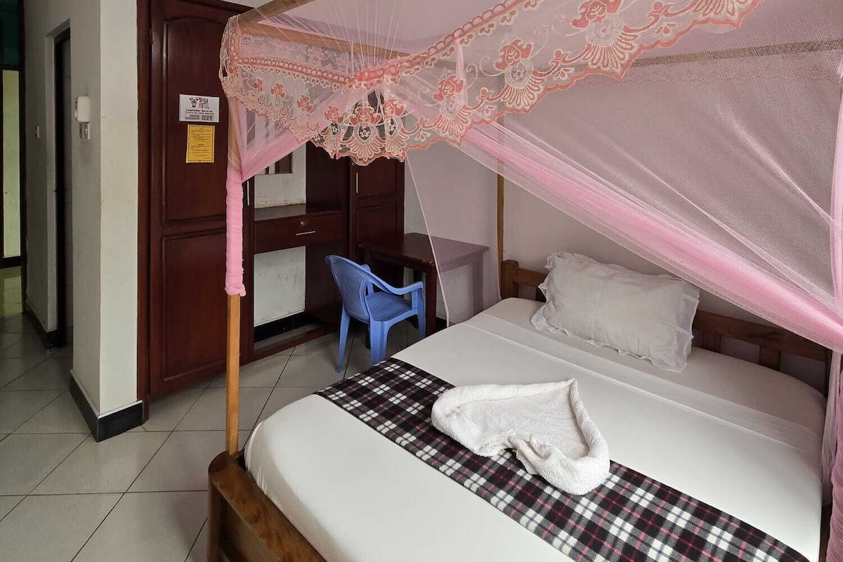 A single bed with a canopy, a desk, a chair, and a ceiling fan in a room with white walls and tiled floor.