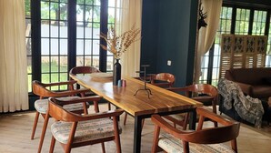 Un espace repas avec une table et des chaises en bois, un luminaire suspendu et une vue sur l’extérieur grâce à de grandes fenêtres.