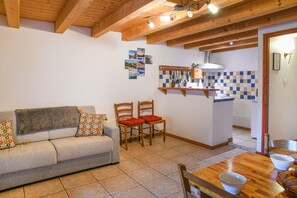 Un salon chaleureux avec un canapé, deux fauteuils, une table à manger en bois et, au fond, une cuisine.