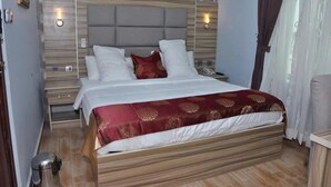 A hotel room with a bed, bedside tables, a nightstand, a trash bin, and a wall-mounted light fixture. - Mozida Suites  (Kano)