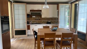 A modern kitchen with a dining area, featuring a large table and chairs, a stove, and a view through the glass door.