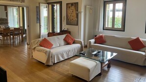 A spacious living room with a sofa, ottoman, and glass coffee table. There is a dining area with a table and chairs, and a kitchen visible through an open door.