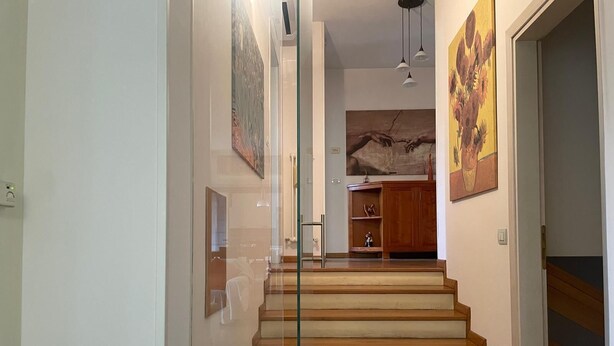 A staircase leading to a landing with a wooden finish, a glass door on the left, and artwork on the walls.