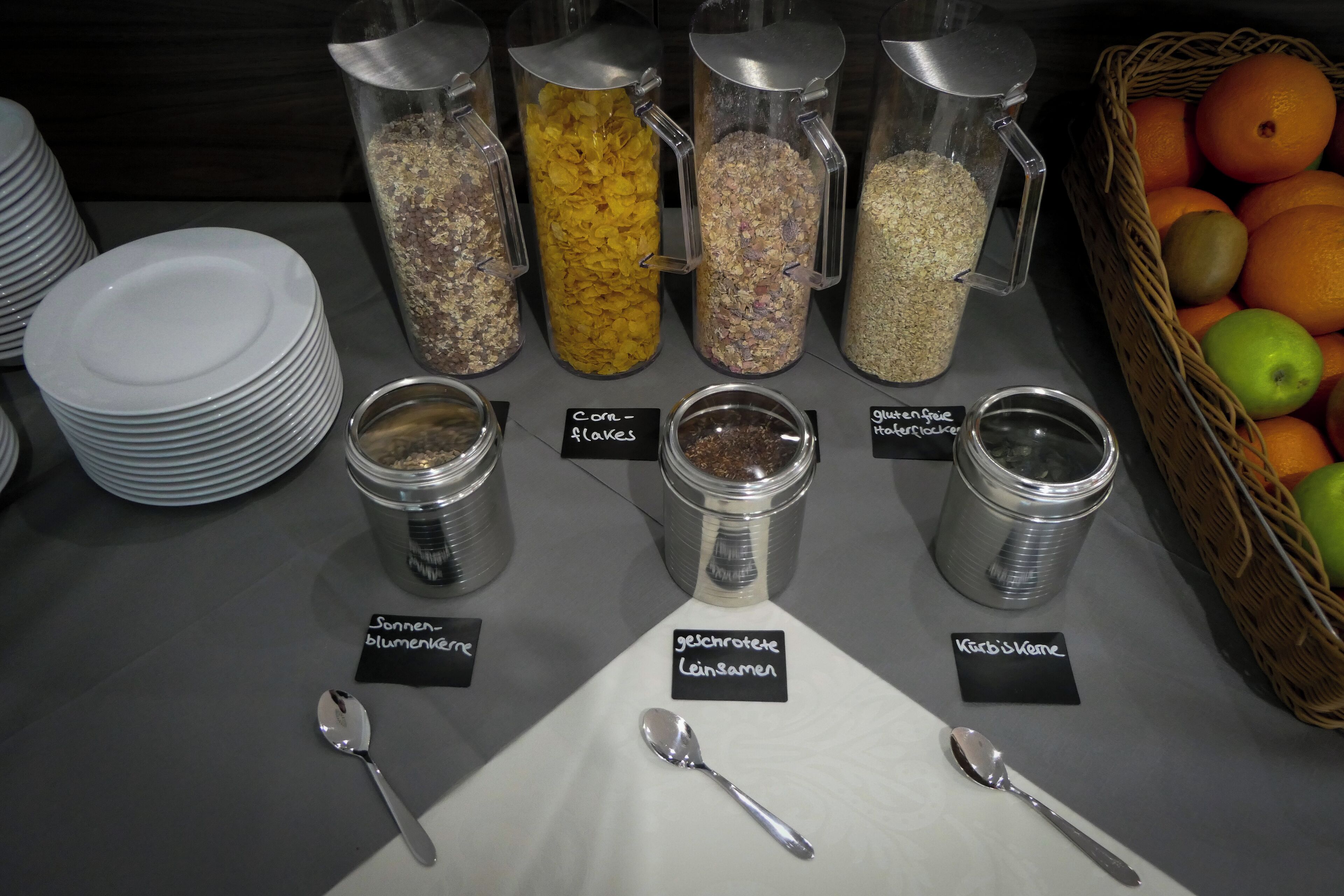 A buffet table with labeled containers of food and a basket of fruits.