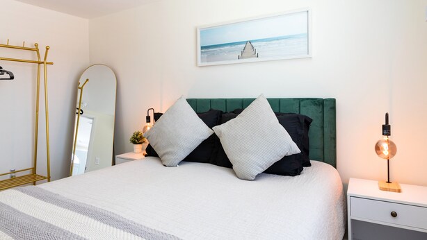 A neatly arranged bedroom with a bed, a bedside table with a lamp, a green upholstered headboard, and a framed picture on the wall.
