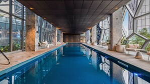 An indoor swimming pool with clear glass walls, modern seating, and a geometric ceiling design. - Designer CBD Executive Suite Heated Pool Gym Sauna (Melbourne)