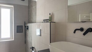 Un bagno moderno con una vasca da bagno ampia, una doccia e un WC. Una finestra permette l'ingresso di luce naturale e su uno scaffale sono presenti articoli da toilette.