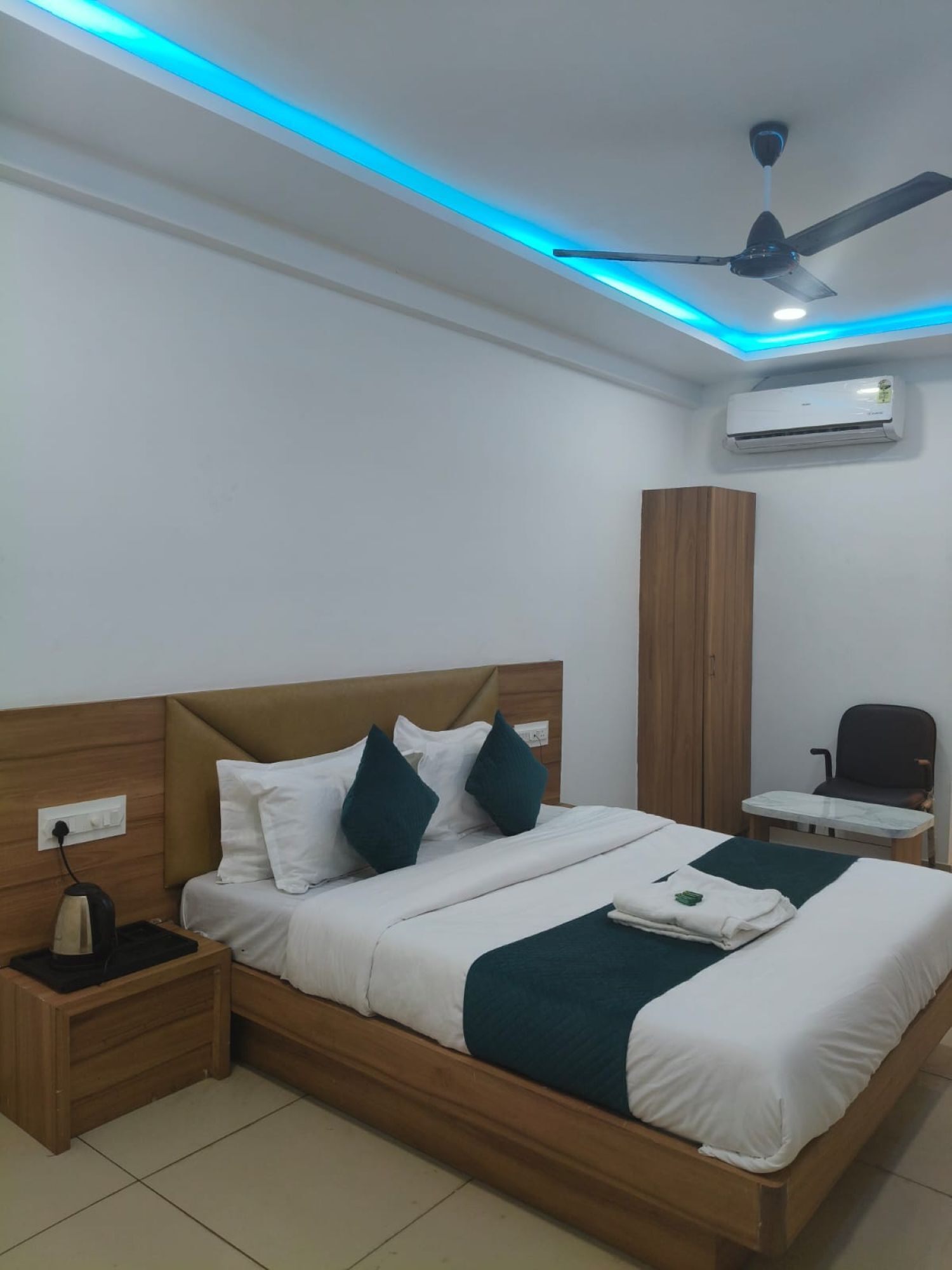 Una habitación de hotel con una cama de madera, ropa de cama blanca, un escritorio con una silla, un equipo de aire acondicionado y un ventilador de techo.