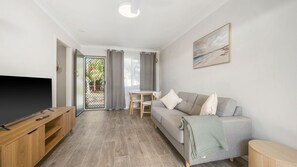 Un saló modern amb televisió de pantalla plana, un sofà gris, un moble de televisió de fusta, una taula de dinar amb cadires i una porta corredissa de vidre que dona a una zona exterior.