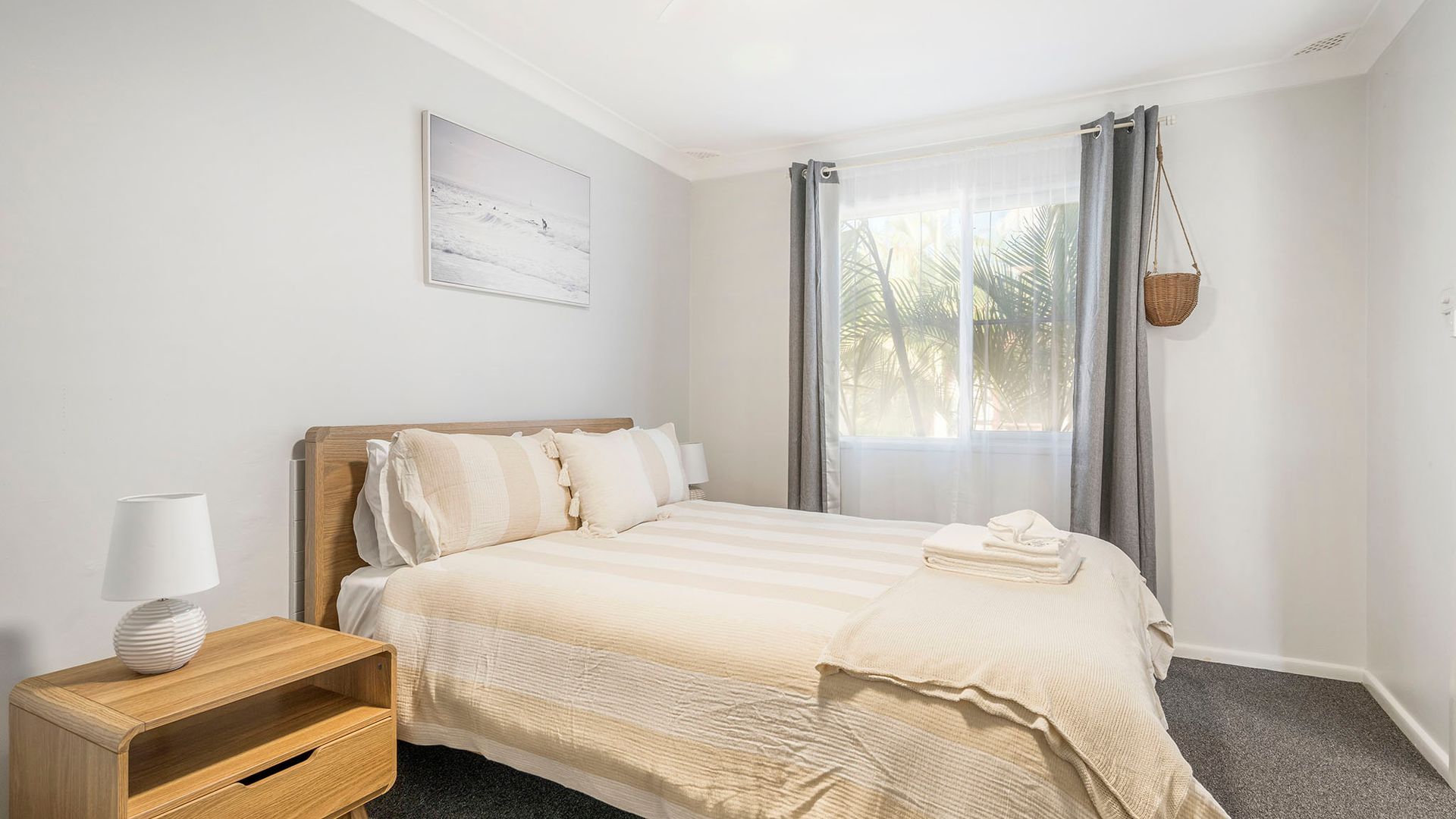 A bedroom with a bed, a bedside table with a lamp, a window with curtains, and a wall-mounted basket.