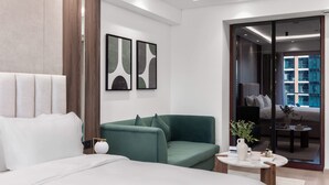 Une chambre d’hôtel moderne avec un grand lit, un canapé vert, une petite table avec un vase, et une vue sur la ville à travers une porte vitrée.