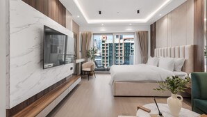 Une chambre moderne avec un grand lit, un téléviseur fixé sur un mur en marbre, un coin salon avec un fauteuil et une vue sur la ville grâce à de grandes fenêtres.
