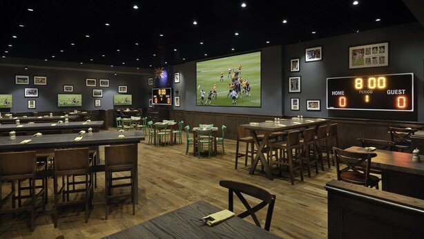 Un bar sportif doté de plusieurs espaces pour s’asseoir, d’un grand écran diffusant un match de football et d’un tableau d’affichage indiquant un match nul à 20h00.