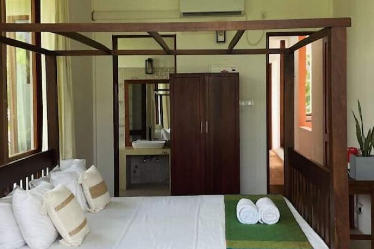 Una cama con dosel y estructura de madera, un ventilador de techo y vista a un baño con lavamanos y espejo.