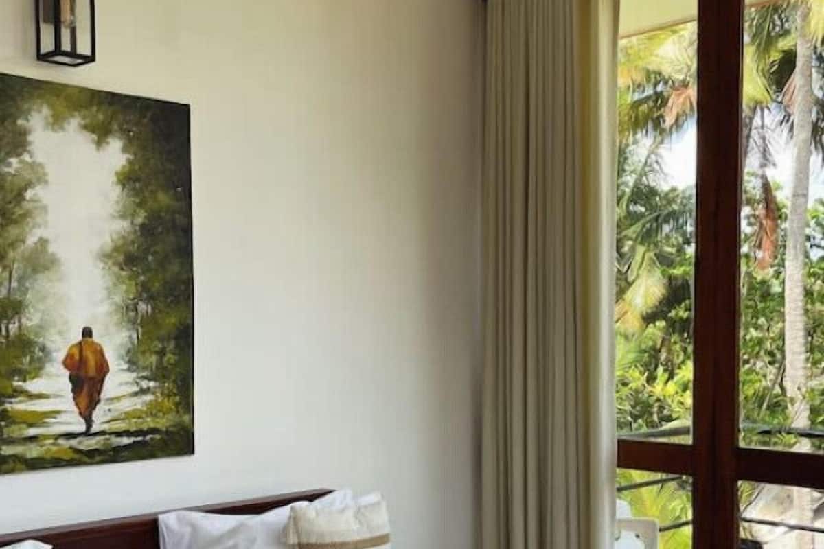Una habitación de hotel con cama, ventilador de techo, un cuadro y vista a follaje por la ventana.