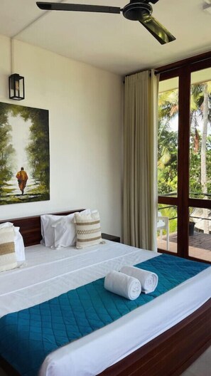 Una habitación de hotel con cama, ventilador de techo, un cuadro y vista a follaje por la ventana.