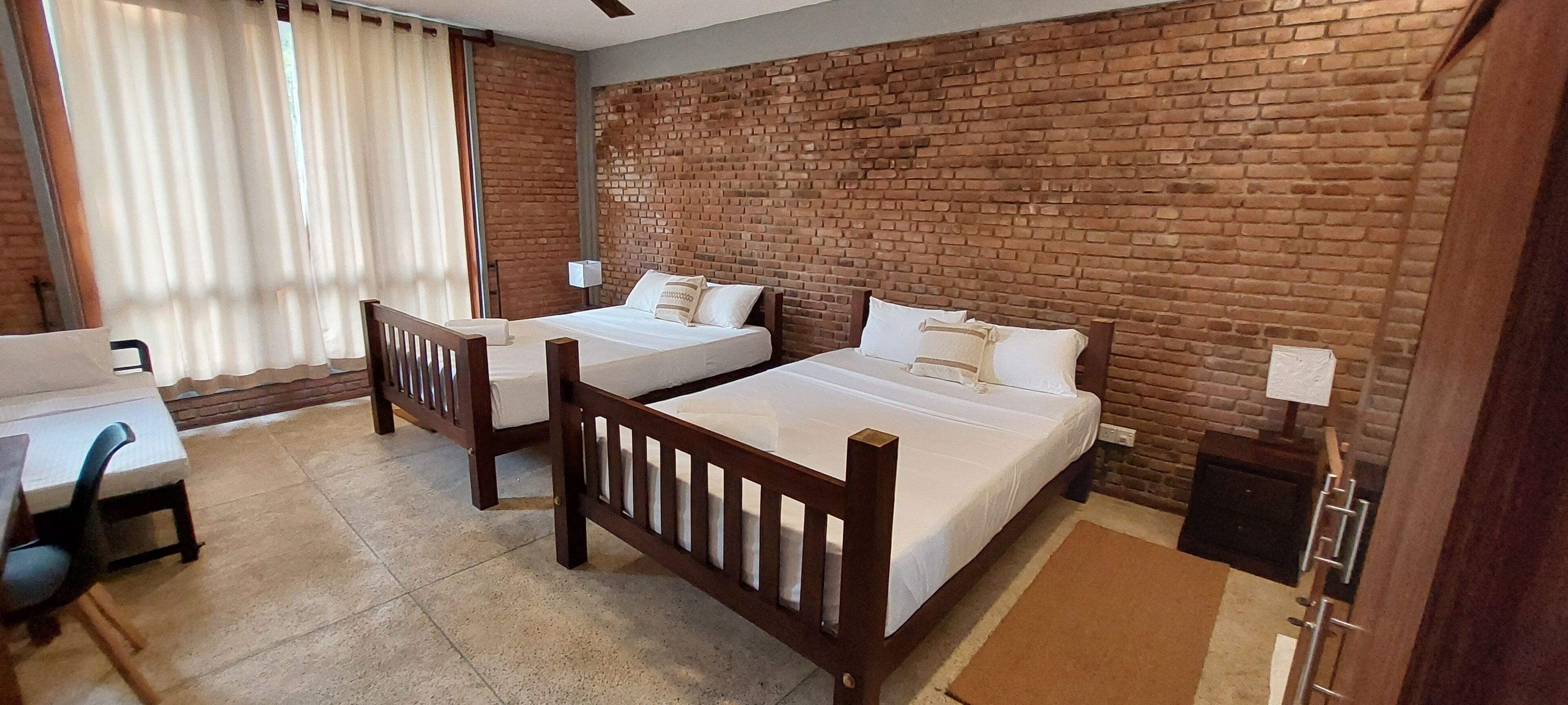 A hotel room with two single beds, a desk, and a chair. The room has a brick wall and a window with curtains.