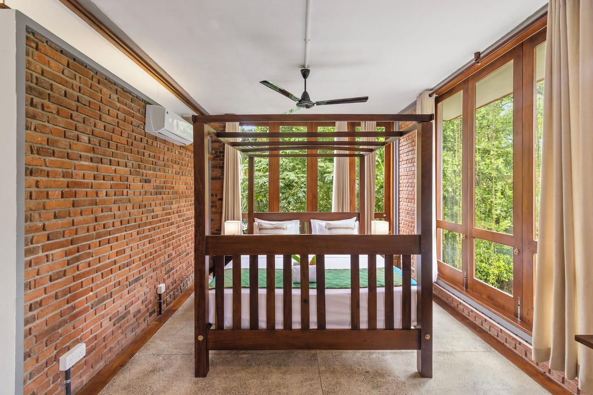 Una habitación con cama de madera, paredes de ladrillo, ventanas amplias y un ventilador de techo.