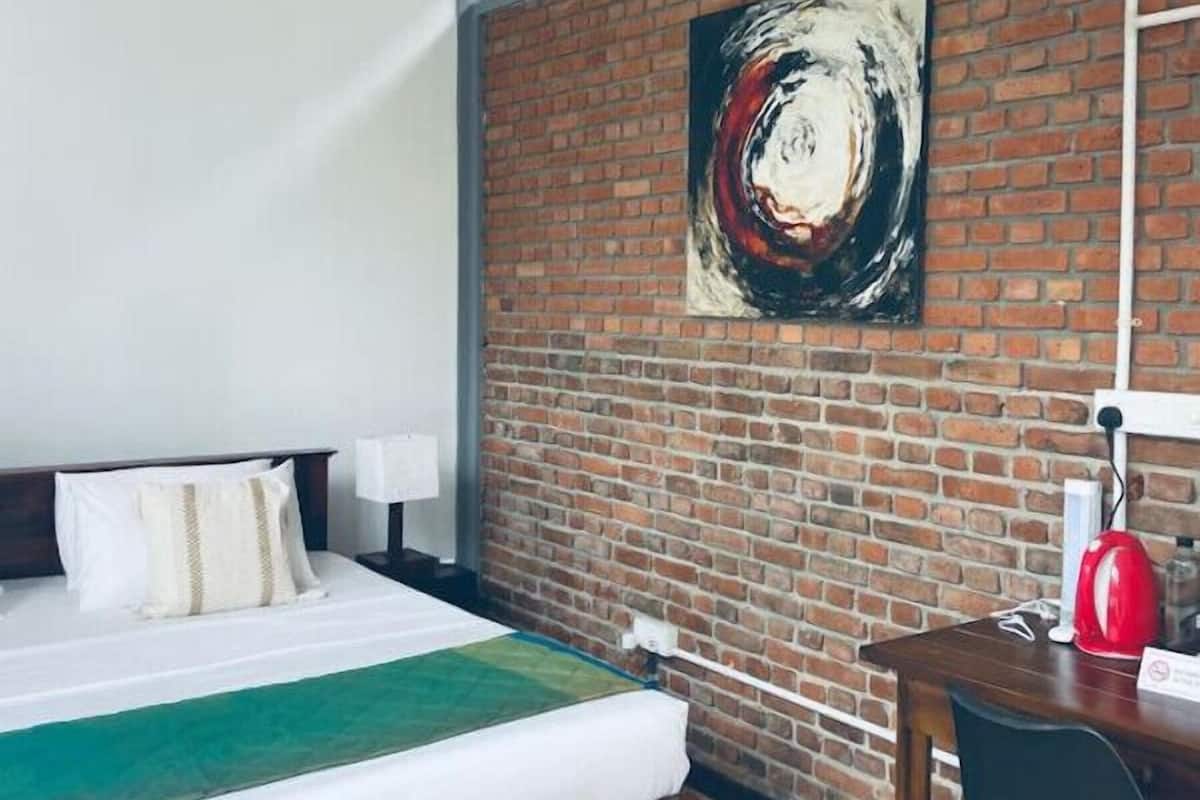 Habitación de hotel con cama, escritorio, lámpara roja y un cuadro abstracto grande sobre una pared de ladrillo.