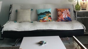 A living room with a white sofa, three cushions, a small turtle on a coffee table, a framed abstract painting, a wall-mounted lamp, and a side table with a vase and lamp. - Charming Apartment in Creole Village of Trois-Ilets (Les Trois-Îlets)
