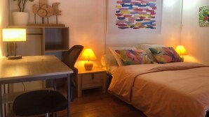 A cozy bedroom with a bed, two bedside lamps, a desk with a chair, a small table, and a wall-mounted shelf with decorative items. - Charming Apartment in Creole Village of Trois-Ilets (Les Trois-Îlets)