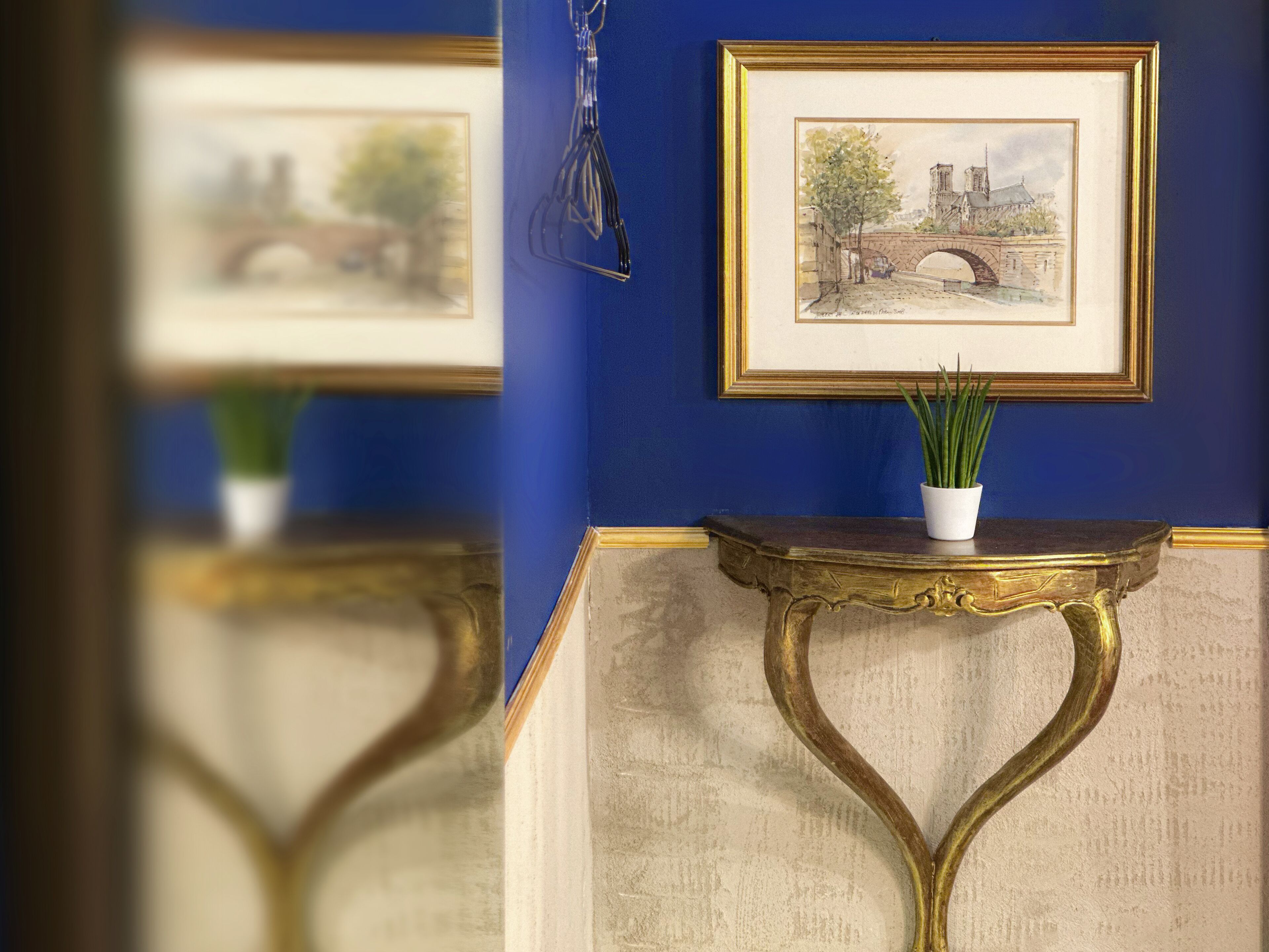 Un quadro ad acquerello incorniciato raffigurante un ponte e un albero su una parete, una piccola vasetto bianco con piante verdi su un tavolo con finiture dorate, e uno specchio che riflette la scena.