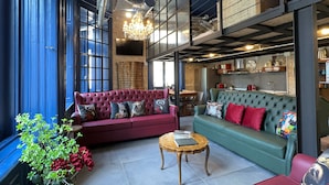 A spacious loft with a red sofa, green sofa, and a wooden coffee table. There is a chandelier hanging from the ceiling, and a kitchen area is visible in the background.