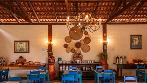 A restaurant interior with blue tables and chairs, a brick wall, a wooden ceiling, and a chandelier. - Pousada Rancho do Ralf (Pirenópolis)