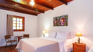 A bedroom with a bed, two bedside tables with lamps, a chair, a small table, and a window with curtains. - Pousada Rancho do Ralf (Pirenópolis)