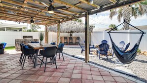A covered patio with a wooden table and chairs, a hanging chair, and a grill area.