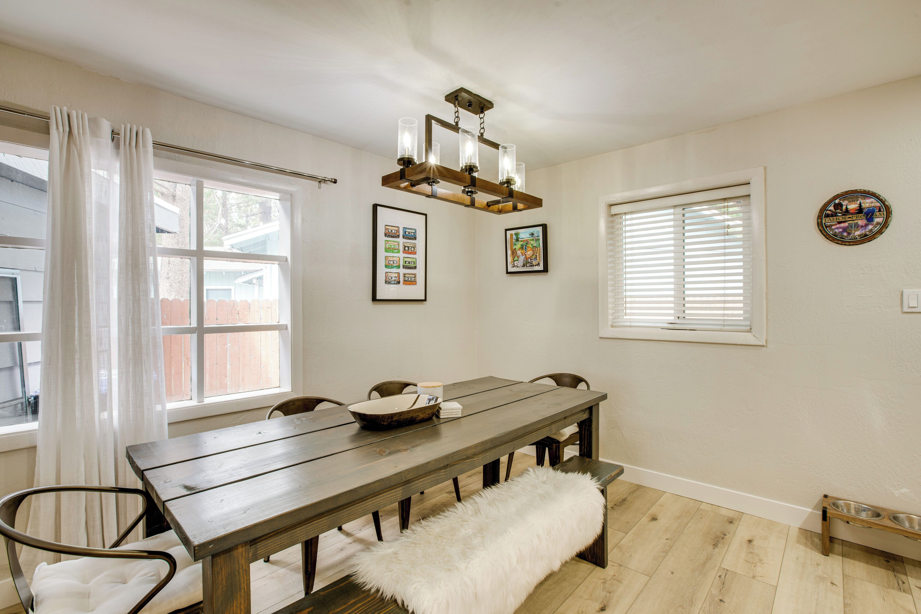 A dining area with a wooden table, chairs, a bench, a chandelier, framed pictures, and a window with blinds.