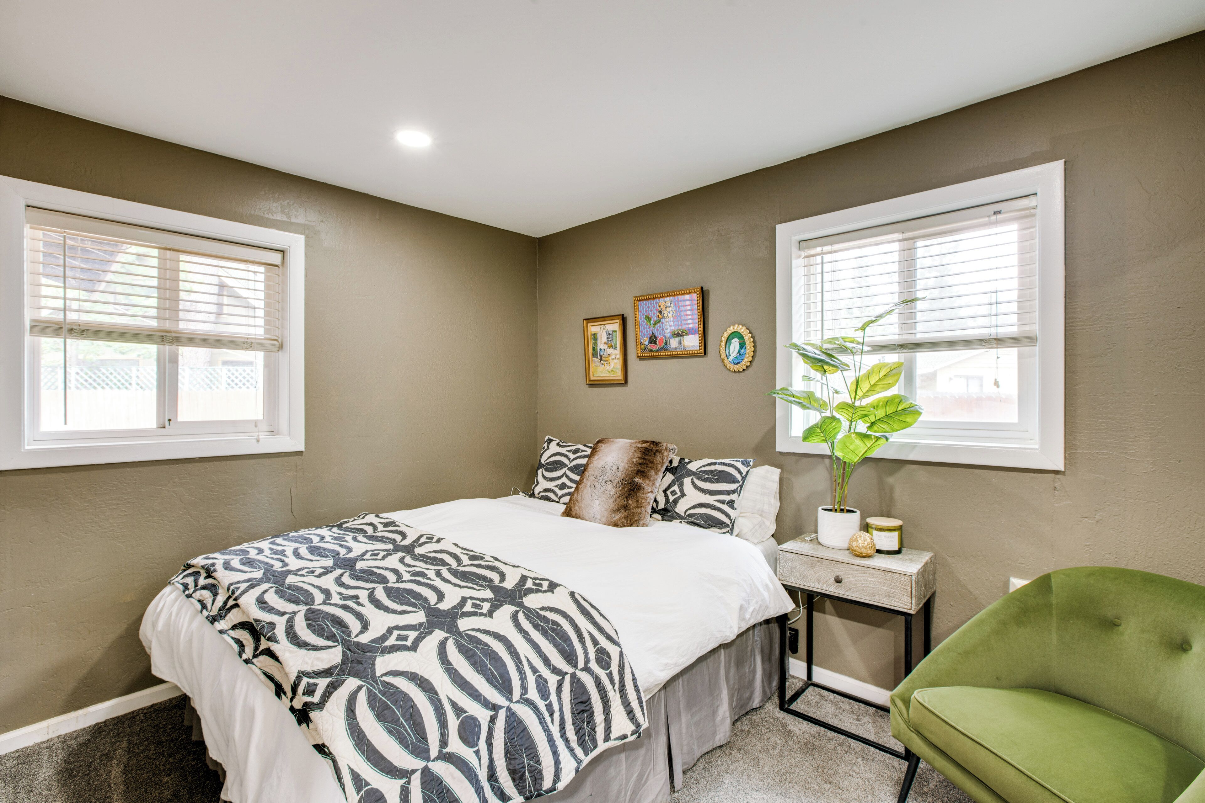 A bedroom with a bed, a green armchair, a bedside table with a plant, and two windows with blinds.