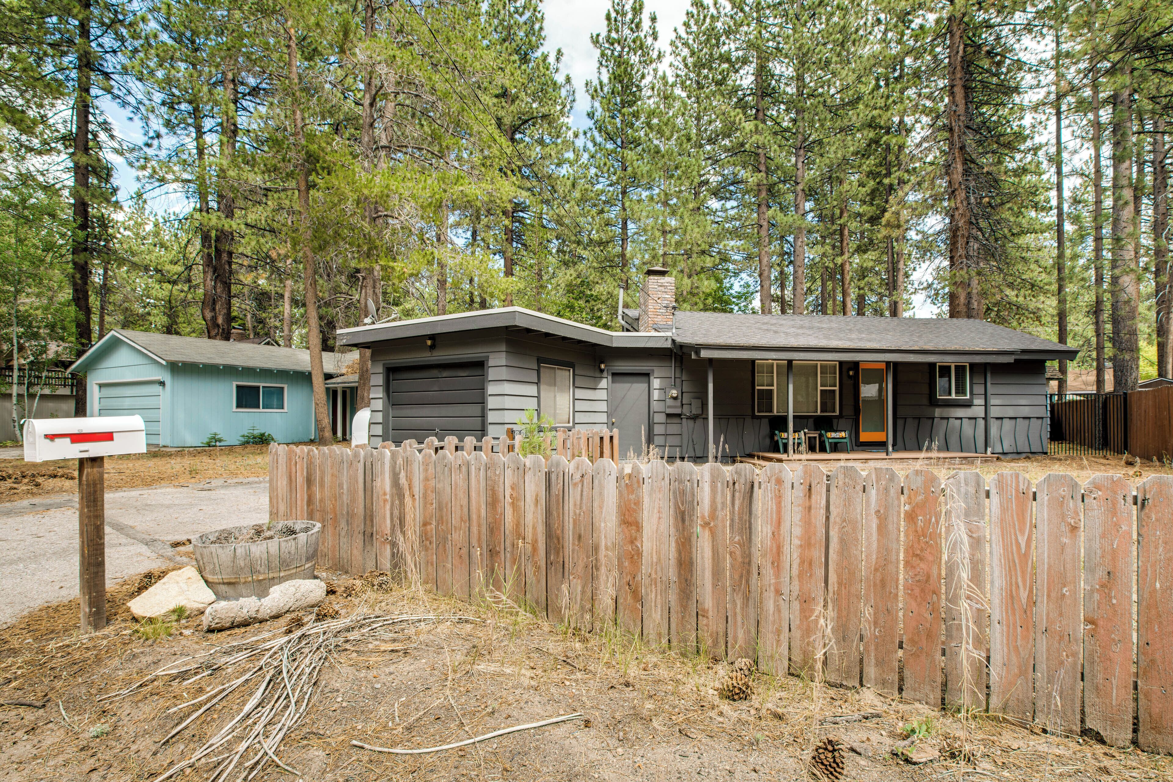A two-story house with a porch, surrounded by a wooden fence and tall pine trees.