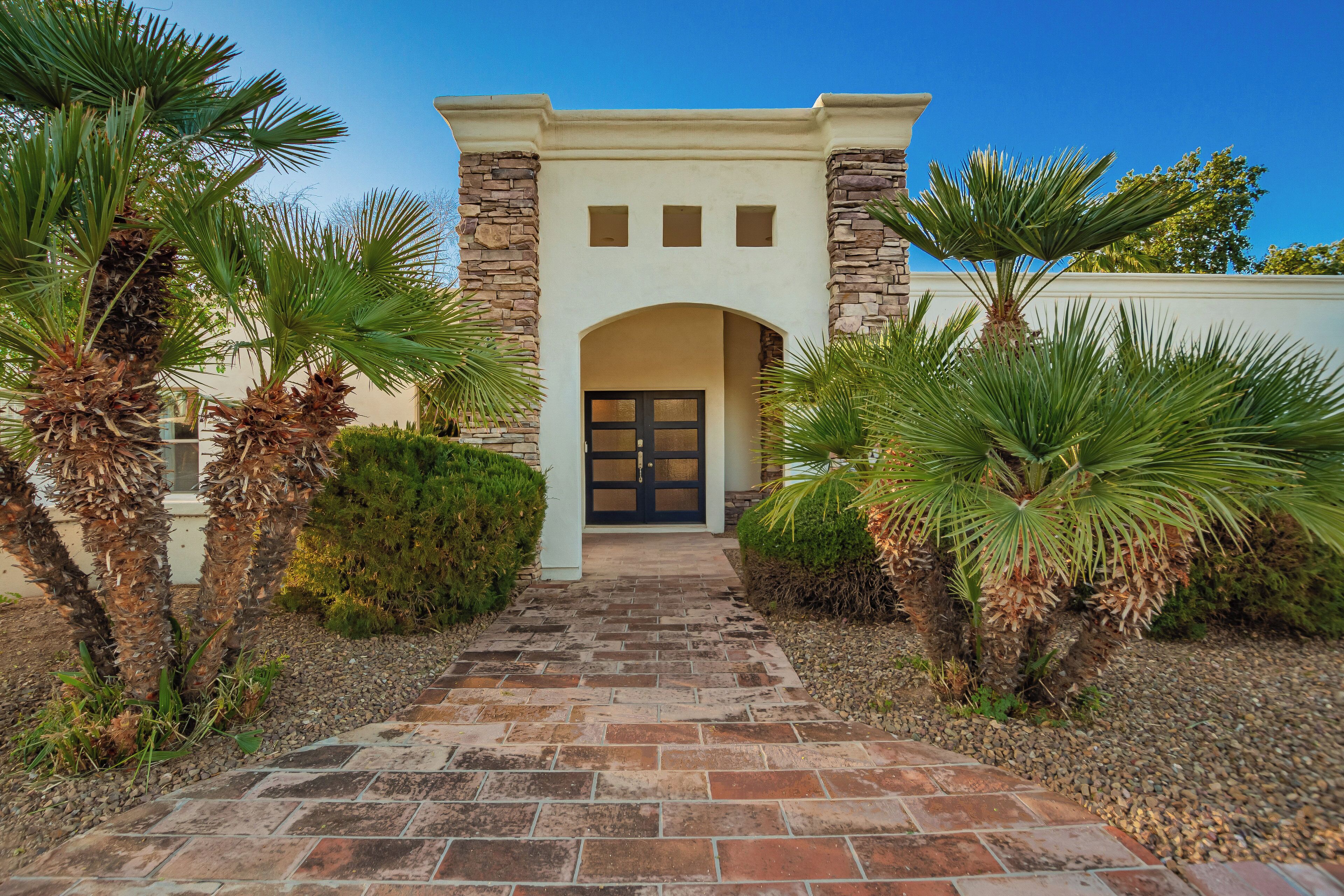 A building entrance with a stone archway, a paved pathway leading to a door, and palm trees on either side.