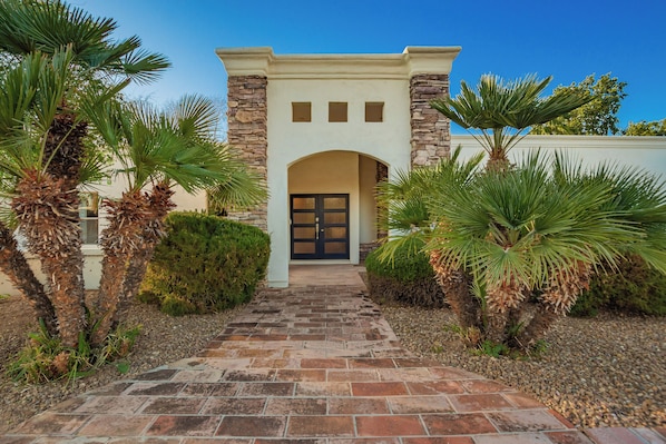 A building entrance with a stone archway, a paved pathway leading to a door, and palm trees on either side.