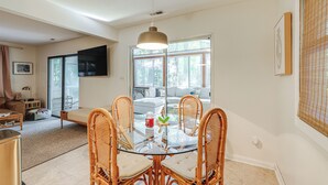 Un comedor con mesa de vidrio y sillas de mimbre, un sofá al fondo y un televisor montado en la pared.