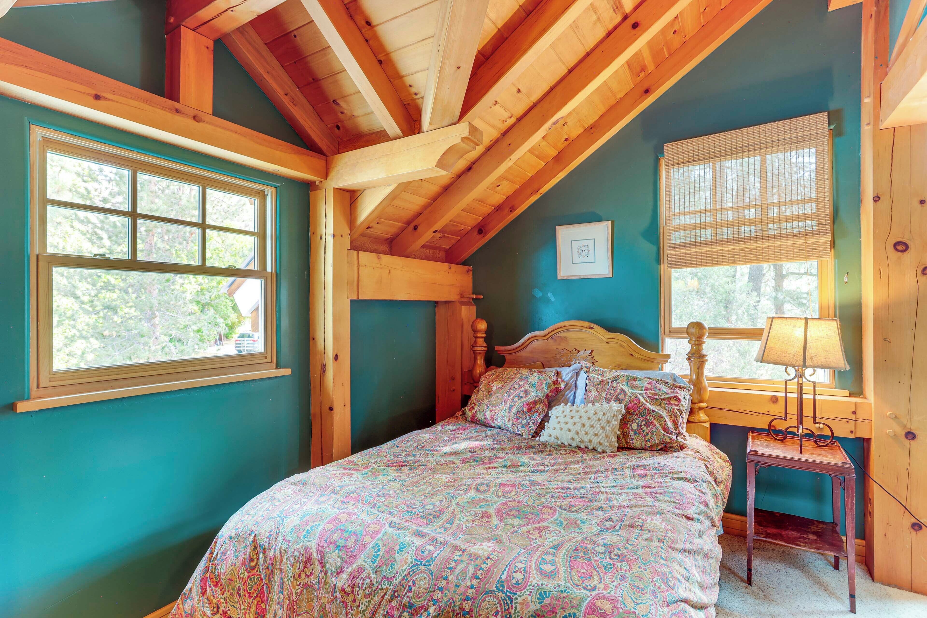 A bedroom with a wooden ceiling, a large bed with patterned bedding, a bedside table with a lamp, and a window with blinds.