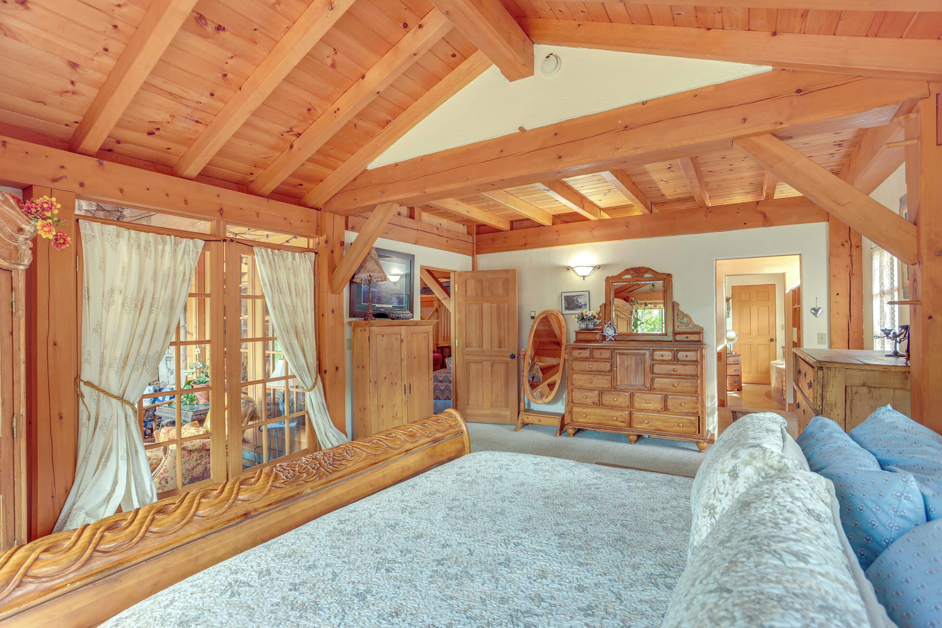 A spacious room with a large bed, wooden ceiling beams, a wooden dresser, and a window with curtains.