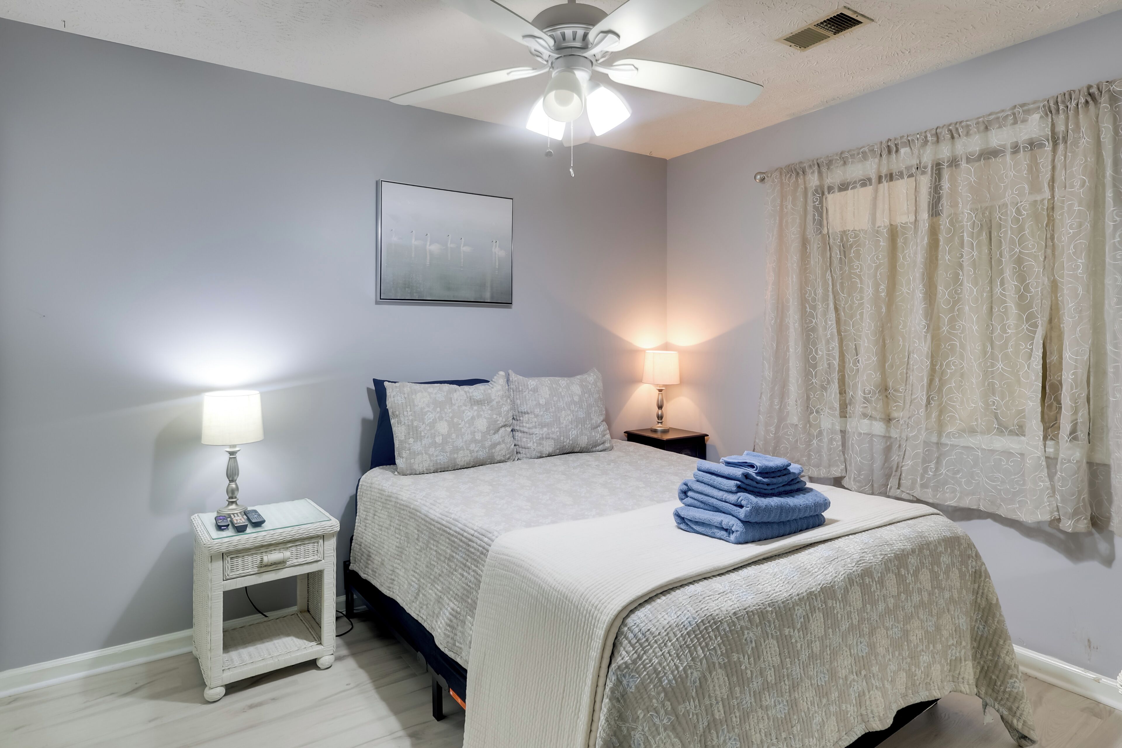 Une chambre à coucher comprenant un lit, une table de chevet, une lampe, un ventilateur de plafond, une fenêtre avec des rideaux et un tableau accroché au mur.