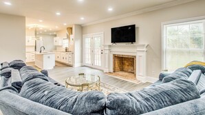 A spacious living room with a fireplace, a television mounted above it, and a modern kitchen in the background. - 3-story Home w/ Private Backyard in Florence! (Florence)