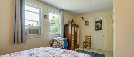 A bedroom with a floral quilted bedspread, a wooden wardrobe, a chair, and a door with a framed picture.