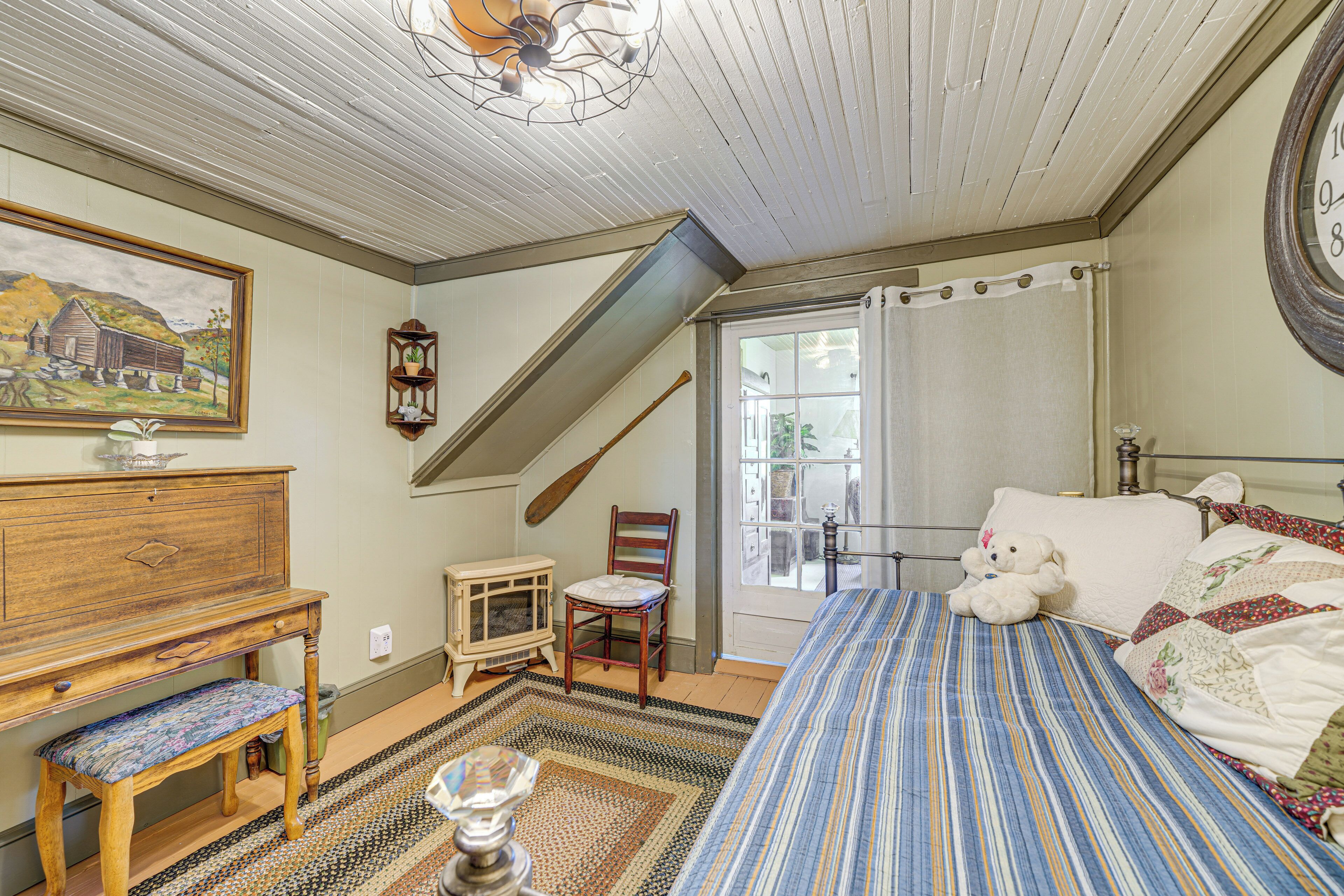 A bedroom with a sloped ceiling, a bed with a striped bedspread, a wooden dresser, a chair, and a window with a view of the outside.