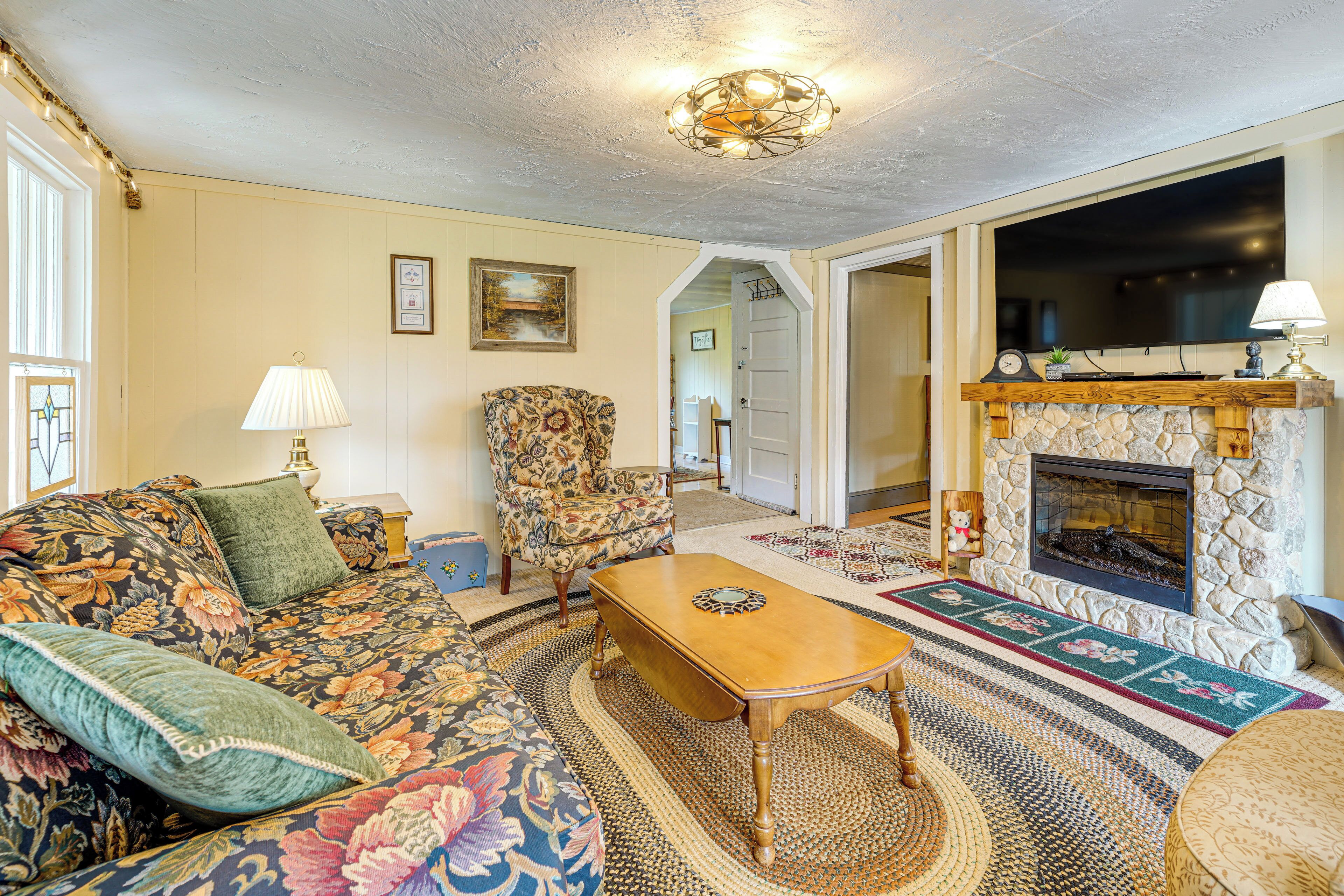 A living room with a floral sofa, a patterned armchair, a wooden coffee table, a fireplace, and a television mounted above it.