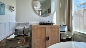 A room with a round mirror, a wooden cabinet with a woven front, a small table with a record player, and a window with curtains. - Walk to Beach: Cottage w/ Yard in Pass Christian! (Pass Christian)
