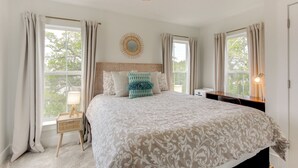 A bedroom with a large bed, a bedside table with a lamp, a window with curtains, and a wall-mounted mirror. - Walk to Beach: Cottage w/ Yard in Pass Christian! (Pass Christian)